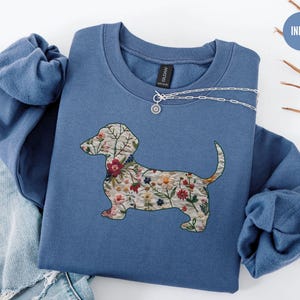Może przedstawiać: Niebieska bluza z nadrukiem w kształcie sylwetki psa rasy dachshund ozdobionej kwiatami. Bluza jest złożona i leży na białym tle. Słowo "INDIGO" jest napisane białym kolorem na niebieskim tle.