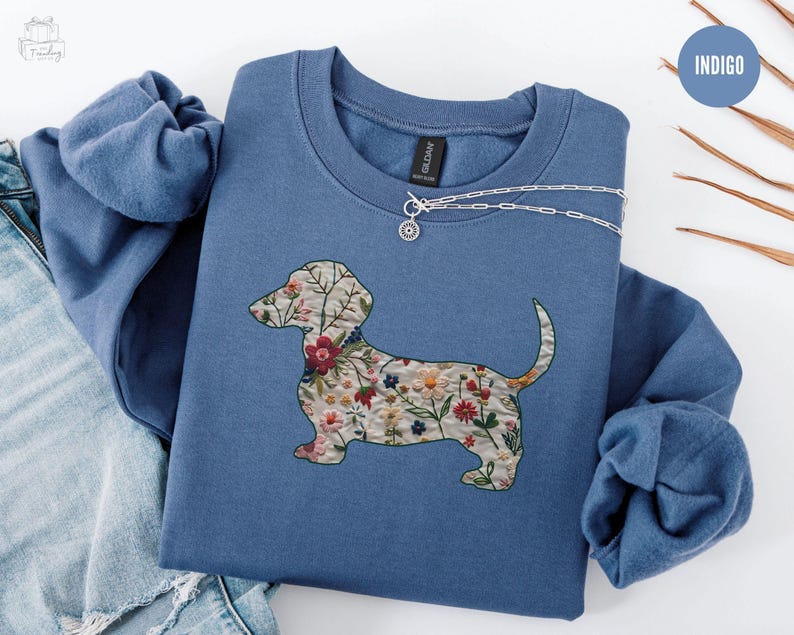 Może przedstawiać: Niebieska bluza z nadrukiem w kształcie sylwetki psa rasy dachshund ozdobionej kwiatami. Bluza jest złożona i leży na białym tle. Słowo "INDIGO" jest napisane białym kolorem na niebieskim tle.