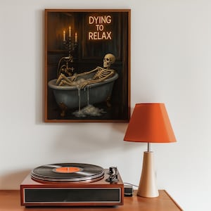 May include: A framed artwork depicts a skeleton relaxing in a bathtub, with the neon text "DYING TO RELAX" above. A vintage record player and a lamp with an orange shade are in the foreground. The artwork is in a wooden frame.