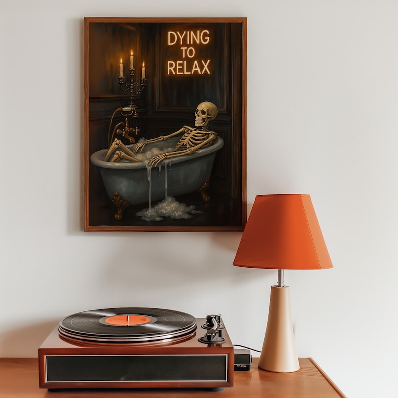 May include: A framed artwork depicts a skeleton relaxing in a bathtub, with the neon text "DYING TO RELAX" above. A vintage record player and a lamp with an orange shade are in the foreground. The artwork is in a wooden frame.
