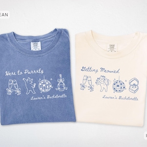 May include: Two t-shirts, one blue and one ivory, with white and blue graphics. The blue shirt says "Here to Purrrty" with cat and party illustrations. The ivory shirt says "Getting Meowied" with similar graphics. Both shirts say "Lauren's Bachelorette".
