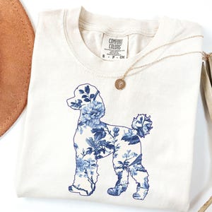 May include: A white t-shirt with a blue floral poodle silhouette design. The shirt is labeled "Comfort Colors" and is available in ivory.