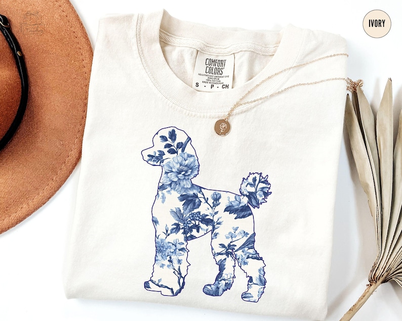 May include: A white t-shirt with a blue floral poodle silhouette design. The shirt is labeled "Comfort Colors" and is available in ivory.