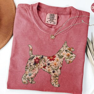 Puede incluir: Una camiseta roja carmes&iacute; con un dise&ntilde;o floral de un perro Schnauzer. La camiseta tiene el texto "COMFORT COLORS" en una etiqueta blanca.
