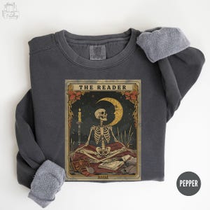 Peut inclure: Sweat-shirt gris foncé avec un motif de carte de tarot représentant un squelette, un croissant de lune et le texte "THE READER". Le motif comprend une bougie, des livres et des éléments floraux. Le sweat-shirt a une doublure en polaire.