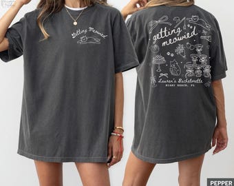 May include: Pepper-colored t-shirt with white text and graphics. The front of the shirt says "Getting Meowied" with a cat graphic. The back of the shirt has the same text, along with a cake, champagne glasses, and "Lauren's Bachelorette, Miami Beach, FL."