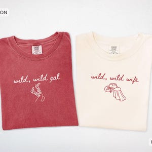 Op de afbeelding: Twee T-shirts met ronde hals. Het karmozijnrode shirt heeft witte tekst "wild, wild gal" met een lijntekening van cowboylaarzen. Het ivoorkleurige shirt heeft donkerrode tekst "wild, wild wife" met een lijntekening van een cowboyhoed.