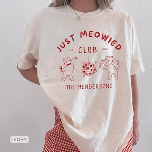 May include: Ivory t-shirt with a red graphic. The text reads "JUST MEOWIED CLUB EST 2026 THE HENDERSONS." The graphic includes cat illustrations, a disco ball, and a bottle. The shirt is a casual, unisex style.