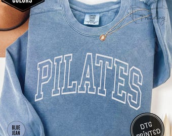 Op de afbeelding: Een spijkerbroekblauwe sweatshirt met het woord "PILATES" in grote witte blokletters. De sweatshirt heeft een ronde hals en lange mouwen. De tekst "COMFORT COLORS" staat in een zwart vakje linksboven. De tekst "BLUE JEAN" staat in een blauwe cirkel linksonder.