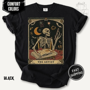 Puede incluir: Camiseta negra con un gr&aacute;fico estilo carta del tarot. El gr&aacute;fico presenta un artista esqueleto con un pincel, una luna creciente y el texto "THE ARTIST". La camiseta tambi&eacute;n tiene el texto "COMFORT COLORS" y "BLACK".