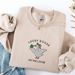 May include: A beige sweatshirt with the text "Lucky Goose on the Loose" and a cartoon goose wearing a green hat and holding a shamrock.