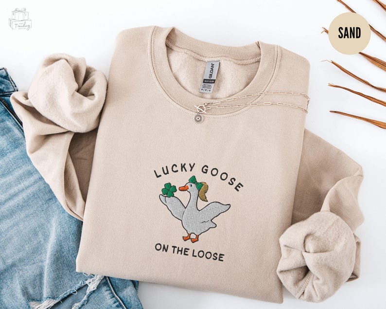 May include: A beige sweatshirt with the text "Lucky Goose on the Loose" and a cartoon goose wearing a green hat and holding a shamrock.