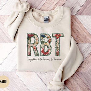 Puede incluir: Una sudadera beige con el texto "RBT Registered Behavior Technician" impreso en la parte delantera. Las letras están delineadas en verde y rellenas con un patrón floral.
