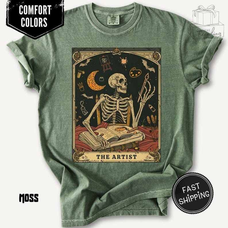 Puede incluir: Camiseta verde musgo con un dise&ntilde;o de carta del tarot. La carta representa a un artista esqueleto con un pincel, sentado frente a un libro abierto, con una luna creciente y &uacute;tiles de arte. El texto "THE ARTIST" est&aacute; en la parte inferior.