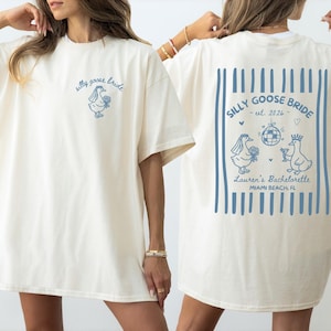 Puede incluir: Una camiseta color marfil con gráficos azules. El frente muestra una pequeña oca con velo y el texto "silly goose bride". La parte posterior presenta un diseño grande con rayas verticales azules, una bola de discoteca, dos ocas y el texto "SILLY GOOSE BRIDE - est. 2026 - Lauren's Bachelorette MIAMI BEACH, FL".