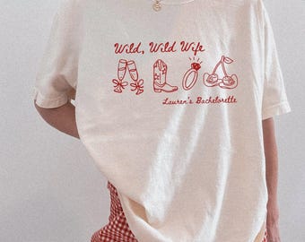 Puede incluir: Camiseta de color marfil con texto y gráficos rojos. El texto dice "Wild, Wild Wife" encima de ilustraciones de copas de champán, una bota de vaquero, un anillo y cerezas. Debajo de los gráficos está escrito "Lauren's Bachelorette".