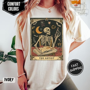 Puede incluir: Camiseta de color marfil con un dise&ntilde;o de carta del tarot de un artista esqueleto. La obra de arte incluye una luna creciente, una paleta de pintura y el texto "THE ARTIST". La camiseta tiene las palabras "COMFORT COLORS" e "IVORY" impresas.