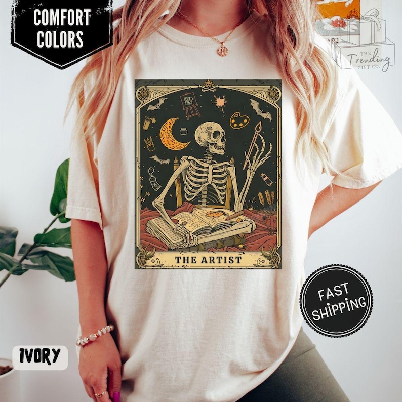 Puede incluir: Camiseta de color marfil con un dise&ntilde;o de carta del tarot de un artista esqueleto. La obra de arte incluye una luna creciente, una paleta de pintura y el texto "THE ARTIST". La camiseta tiene las palabras "COMFORT COLORS" e "IVORY" impresas.