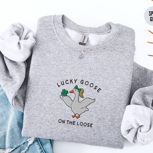 May include: A gray sweatshirt with the text "Lucky Goose on the Loose" and a cartoon goose wearing a green hat and holding a shamrock.
