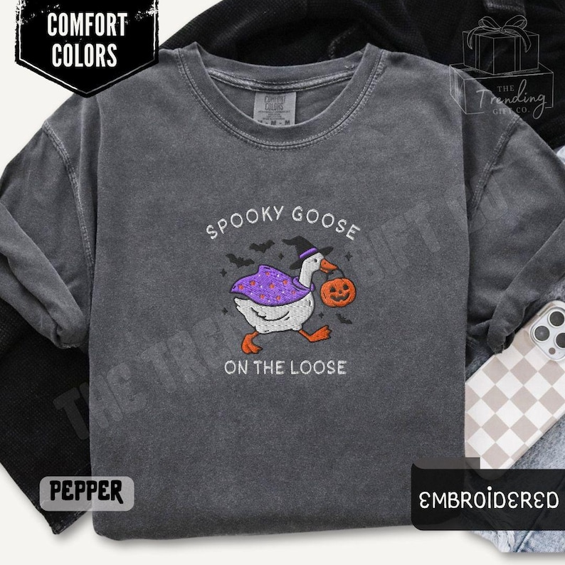May include: Pepper-colored, long-sleeved shirt with an embroidered design. The design features a goose wearing a witch hat and carrying a jack-o'-lantern, with the text "Spooky Goose on the Loose". The shirt is labeled "Comfort Colors" and "Embroidered".
