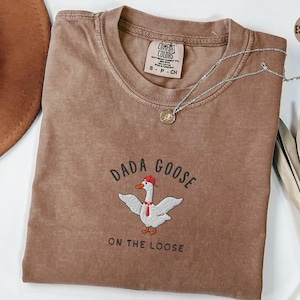 May include: A brown t-shirt with the text "Dada Goose on the Loose" and an embroidered image of a goose wearing a red hat. The shirt is made of a soft, comfortable material.