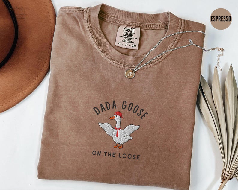 May include: A brown t-shirt with the text "Dada Goose on the Loose" and an embroidered image of a goose wearing a red hat. The shirt is made of a soft, comfortable material.