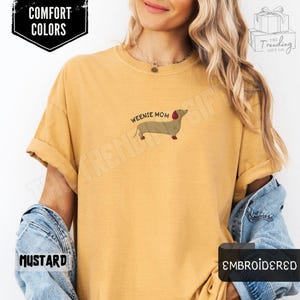 May include: Mustard-colored t-shirt with the text "Weenie Mom" and an embroidered dachshund dog. The shirt also features the words "Comfort Colors" and "Embroidered".