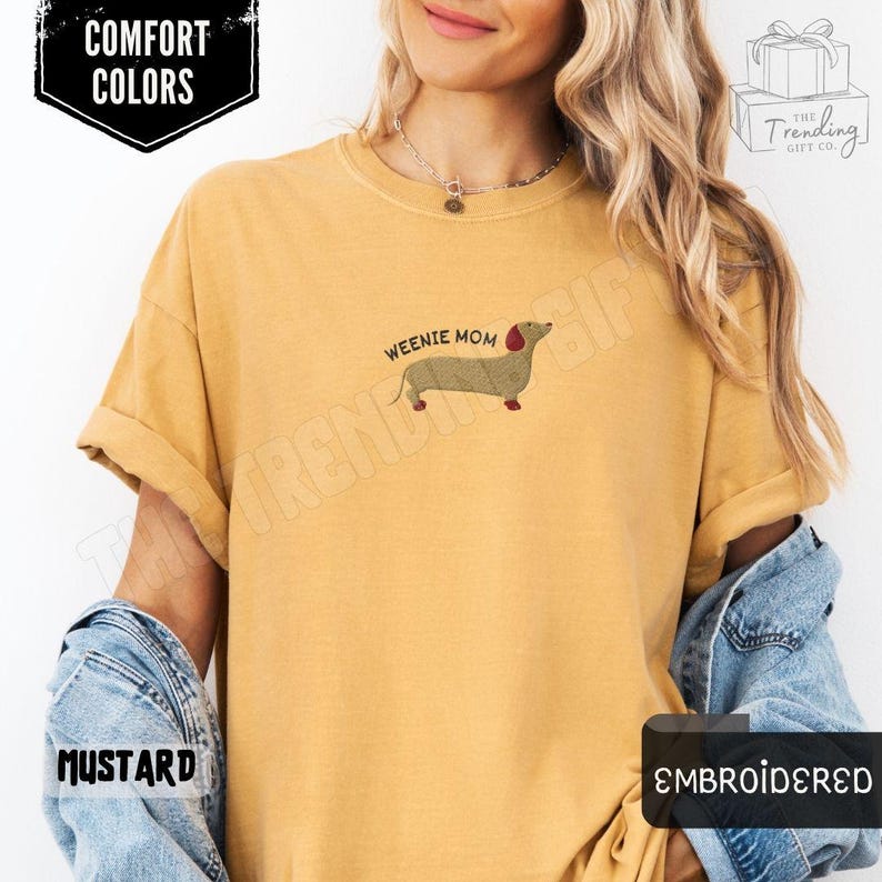 May include: Mustard-colored t-shirt with the text "Weenie Mom" and an embroidered dachshund dog. The shirt also features the words "Comfort Colors" and "Embroidered".