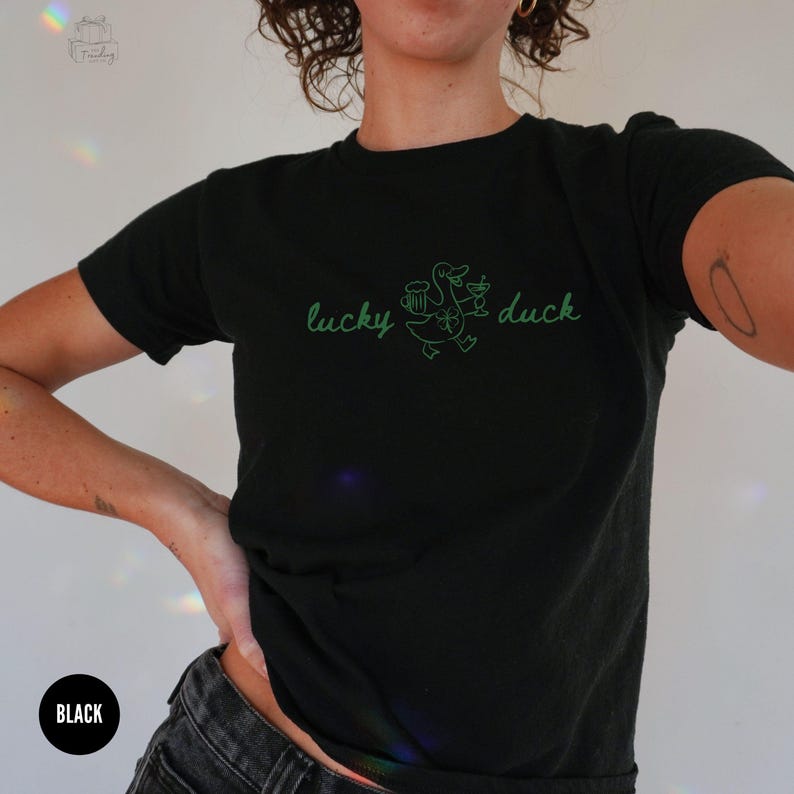 Op de afbeelding: Zwart T-shirt met de tekst "lucky duck" in groen schrift. Een groene cartoon eend met een bierpul en een cocktailglas staat gecentreerd tussen de woorden. Het shirt heeft korte mouwen.
