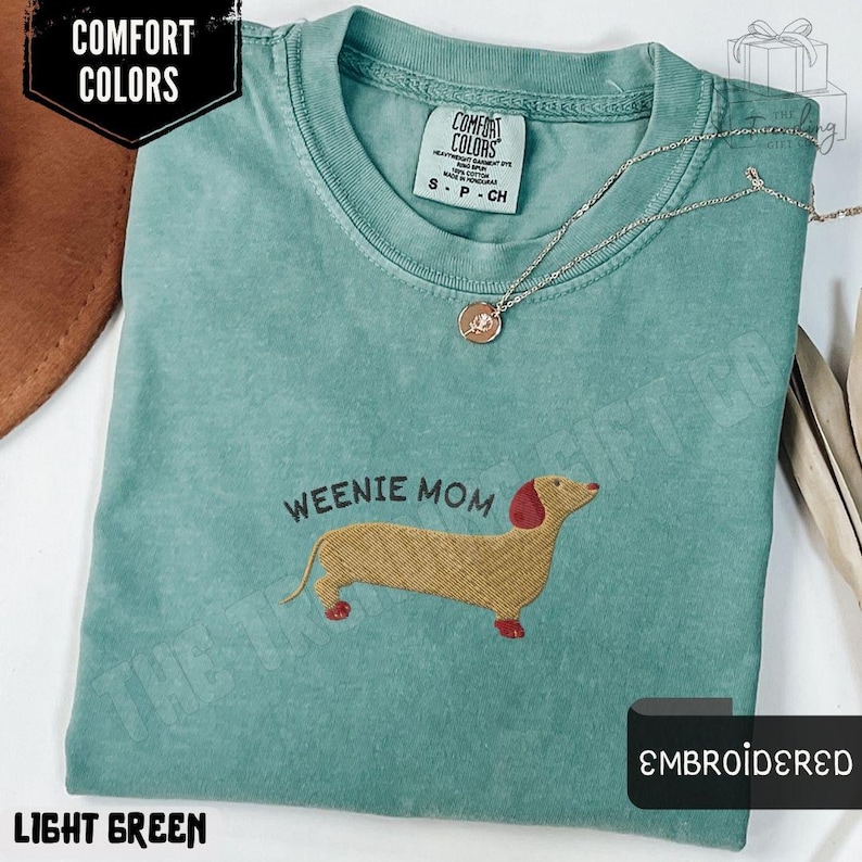 May include: Light green t-shirt with the embroidered text "WEENIE MOM" and a dachshund design. The shirt has a crew neck and short sleeves. The shirt is labeled "Comfort Colors" and is a size small.