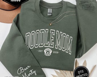 Puede incluir: Sudadera verde militar con texto blanco "DOODLE MOM" y un gráfico de perro doodle. La manga muestra "Bear Morty". La sudadera es una Gildan 18000. También es visible el texto "EST 2026".
