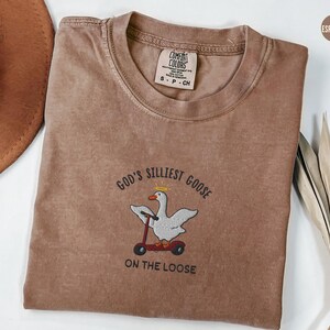 May include: A brown t-shirt with the text "God's Silliest Goose On The Loose" and an embroidered image of a goose riding a scooter.