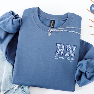 Könnte beinhalten: Ein blaues Sweatshirt mit einem weißen gestickten Design, das "RN Emily" sagt.