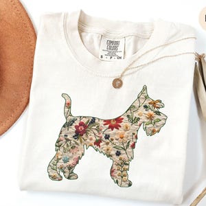 Puede incluir: Una camiseta blanca con un dise&ntilde;o floral en forma de perro. El dise&ntilde;o est&aacute; en tonos de rojo, rosa, amarillo y verde. La camiseta tiene una etiqueta que dice "Comfort Colors" y el texto "S - P - CH". La camiseta est&aacute; sobre un fondo blanco con un sombrero marr&oacute;n y una planta seca.
