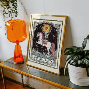 Puede incluir: Una impresi&oacute;n enmarcada en dorado de una carta de tarot con un esqueleto montando un unicornio arco&iacute;ris. La carta se titula "The Audacity".