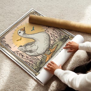 May include: A vintage-style illustration of a white goose with a yellow beak and feet, standing in a field of green grass and flowers. The goose is surrounded by a decorative frame with a light pink background.
