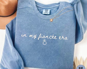 May include: A blue jean-colored sweatshirt with the phrase "in my fiancee era" embroidered in white script, along with a ring graphic. A gold necklace with a pendant rests on the shirt. A brown hat is visible in the background.