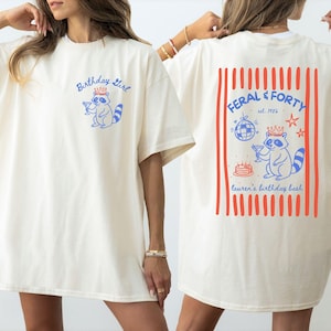 以下が含まれることがあります： アイボリー色のTシャツで、胸元には王冠を被りカクテルを持った青いアライグマのグラフィックと「Birthday Girl」の文字があります。背面には赤いストライプのデザインがあり、アライグマ、ミラーボール、ケーキ、そして「Feral & Forty est. 1986」という文字が描かれています。
