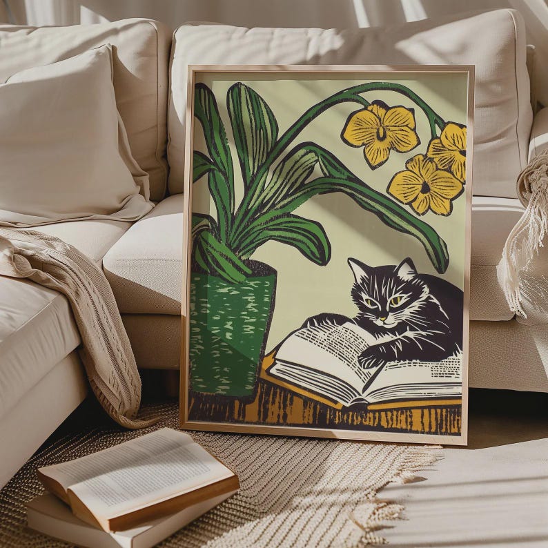 May include: A framed print of a black and white cat reading a book. The cat is sitting on a table with a green potted plant with yellow flowers behind it.