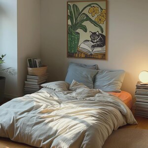 May include: A white bed with a light beige duvet cover and two blue pillows. A framed print of a black cat reading a book with a yellow flower behind it hangs on the wall above the bed.