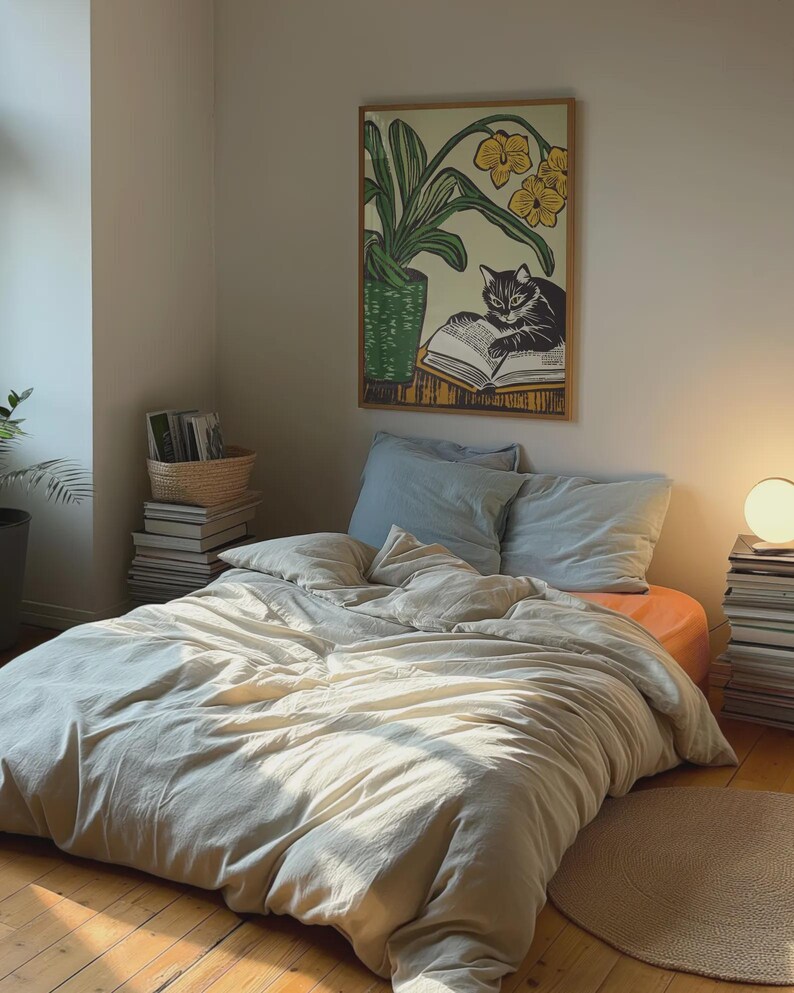May include: A white bed with a light beige duvet cover and two blue pillows. A framed print of a black cat reading a book with a yellow flower behind it hangs on the wall above the bed.