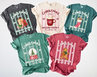 May include: Five short-sleeve t-shirts in charcoal, cream, teal, green, and red. Each features a vertical striped background with the text CHRISTMAS CREW 2026, WILSON FAMILY, and New York, N.Y. Graphics include gifts, a cocoa mug, stocking, snow globe, and gingerbread man.