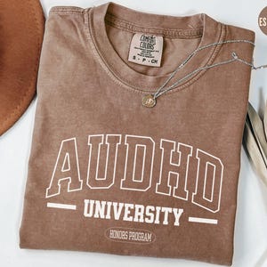 May include: A brown t-shirt with the text "ADHD University - Honors Program" in white. The shirt is a relaxed fit and has a distressed look.