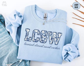 May include: Light blue sweatshirt with the letters LCSW in a floral design. The words "licensed clinical social worker" are printed below the letters. A silver necklace with a small pendant is on the sweatshirt.
