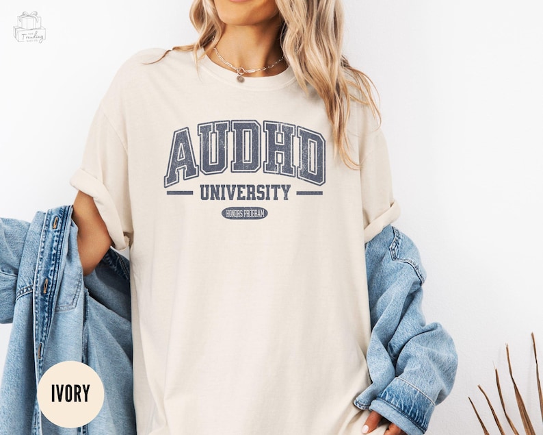 May include: A beige t-shirt with a blue graphic that reads "AUDHD University - Honours Program".