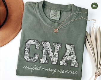 Könnte beinhalten: Ein grünes T-Shirt mit einem floralen Druck, der "CNA" buchstabiert und den Text "certified nursing assistant" trägt.