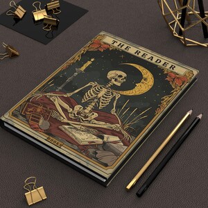 May include: A hardcover journal featuring a skeleton seated before a crescent moon and starry night sky. The journal cover reads "THE READER" and includes a candle, books, and floral accents. Gold and black pencils and binder clips are also present.