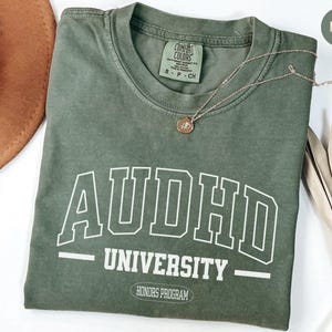 May include: A green t-shirt with the text "AUDHD UNIVERSITY - HONORS PROGRAM" in white. The shirt is made of a soft, comfortable material and has a vintage look.