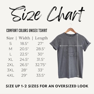 May include: A size chart for a Comfort Colors unisex t-shirt, with measurements in inches. The chart includes sizes S to 4XL, with width and length dimensions. The image also shows a gray t-shirt on a hanger.