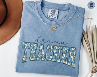 Puede incluir: Una camiseta azul vaquera Comfort Colors con las palabras "drama TEACHER" en un diseño floral. Un collar dorado con un colgante descansa sobre la camisa. La camisa está doblada y colocada sobre una superficie con un sombrero marrón y plantas secas.